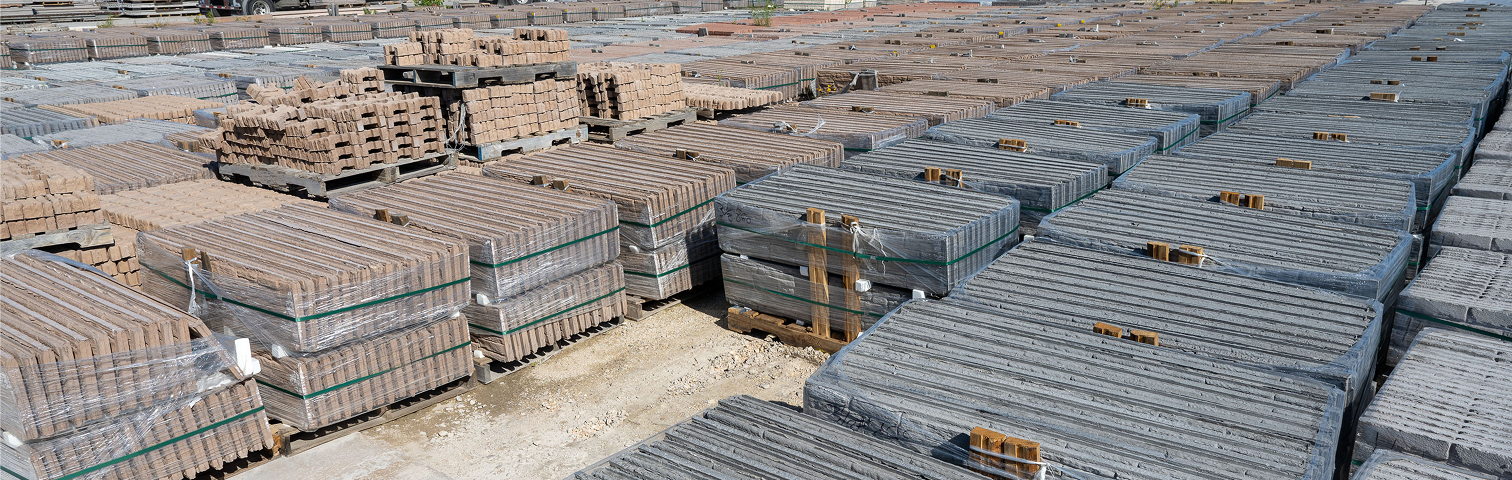 Rows of stacked pallets filled with bricks and concrete blocks are arranged in an open yard, ready for transport or construction use. The materials are neatly bundled and partially wrapped in plastic.