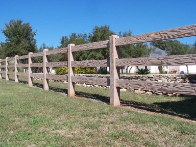 A close-up view of a wooden ranch-style fence with three horizontal rails, standing on a grassy lawn with trees and a stone wall in the background under a clear blue sky.