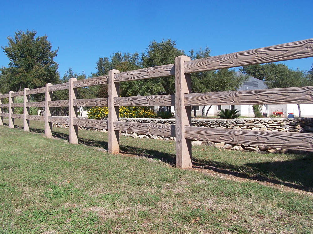 A brown wooden ranch-style fence runs alongside a grassy yard, with trees and a stone wall visible in the background under a clear blue sky.