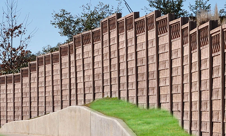 A tall brown concrete sound barrier wall with a patterned design runs alongside a road, with trees visible behind the wall and green grass growing near its base.