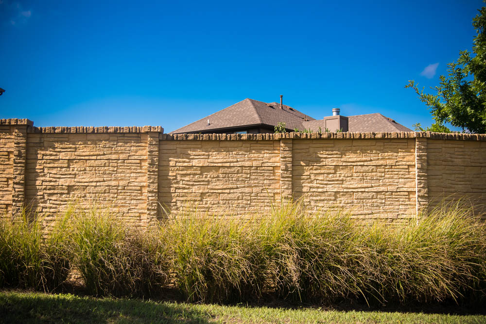 Tan stone wall with tall grass in front, set against a clear blue sky. Rooftops of houses and part of a tree are visible behind the wall.