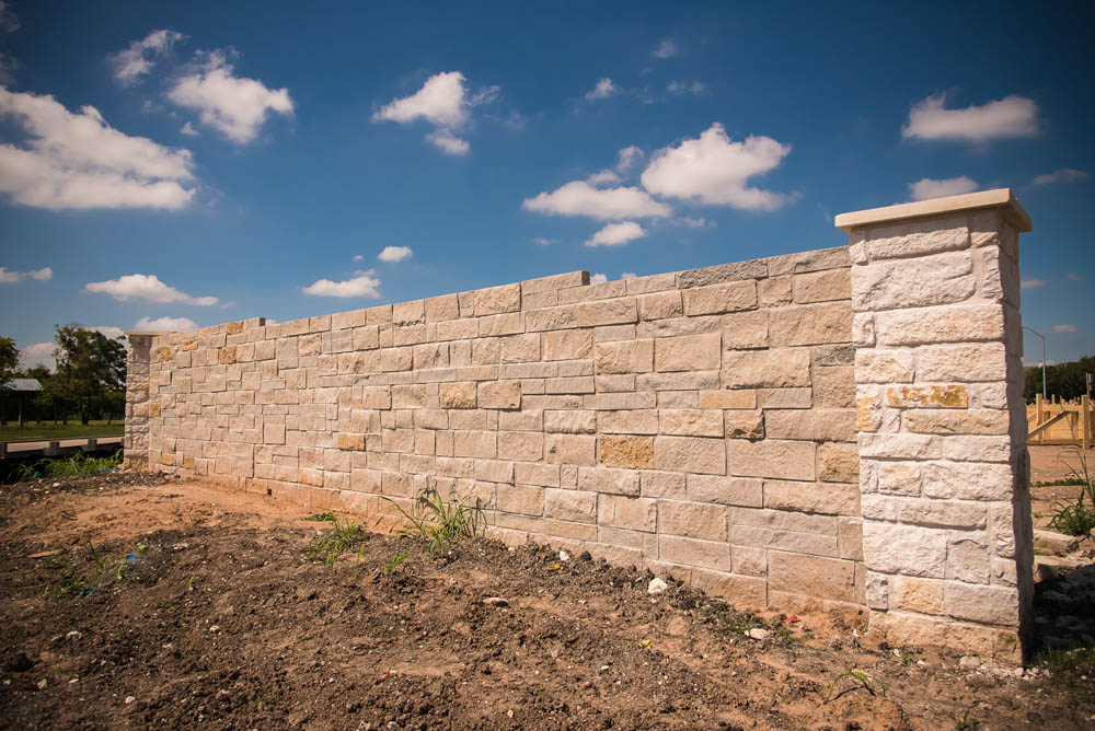 A tall stone wall with light-colored bricks stands on bare soil under a blue sky with scattered clouds. Sparse grass and small plants grow at the base of the wall.