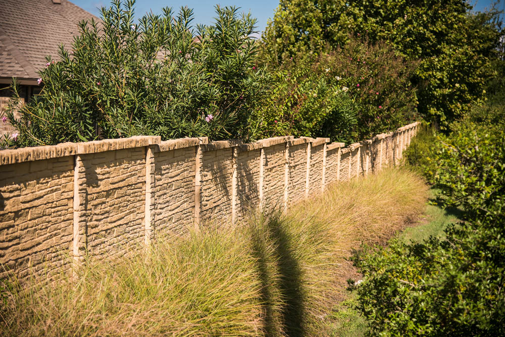 A beige stone backyard fence with vertical posts runs alongside tall grass, leafy bushes, and trees under a sunny blue sky.