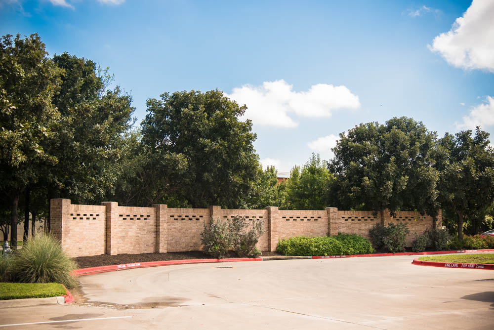 A tan brick wall with decorative rectangular cutouts runs along the edge of a parking lot, bordered by trees and shrubs under a blue sky with scattered clouds.