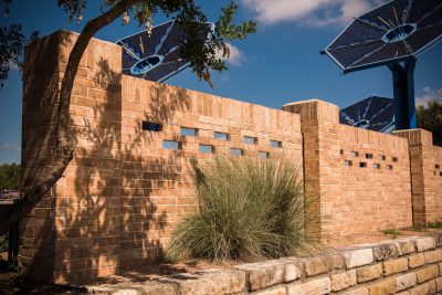 A brick wall with rectangular glass blocks and tall grasses in front, partially shaded by a tree, with large blue solar panel structures visible in the background against a blue sky.