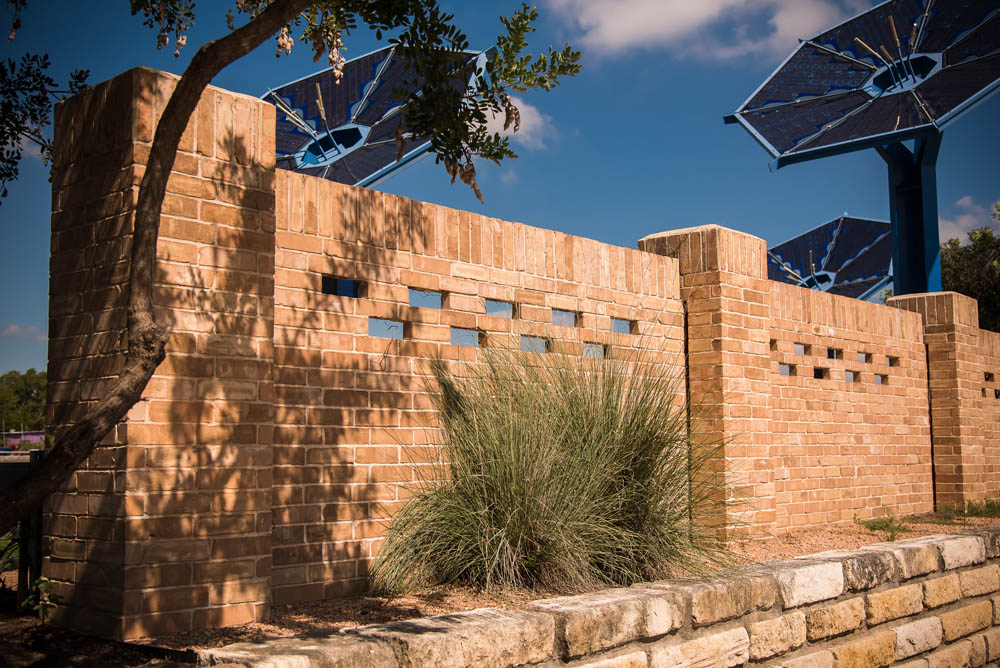 A brick wall with rectangular glass blocks and tall grasses in front, partially shaded by a tree, with large blue solar panel structures visible in the background against a blue sky.