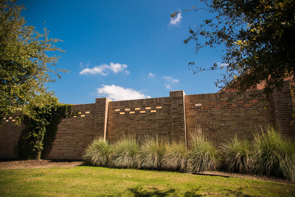 A tall brick wall with decorative rectangular openings, surrounded by green grass, shrubs, and trees under a bright blue sky with a few clouds.