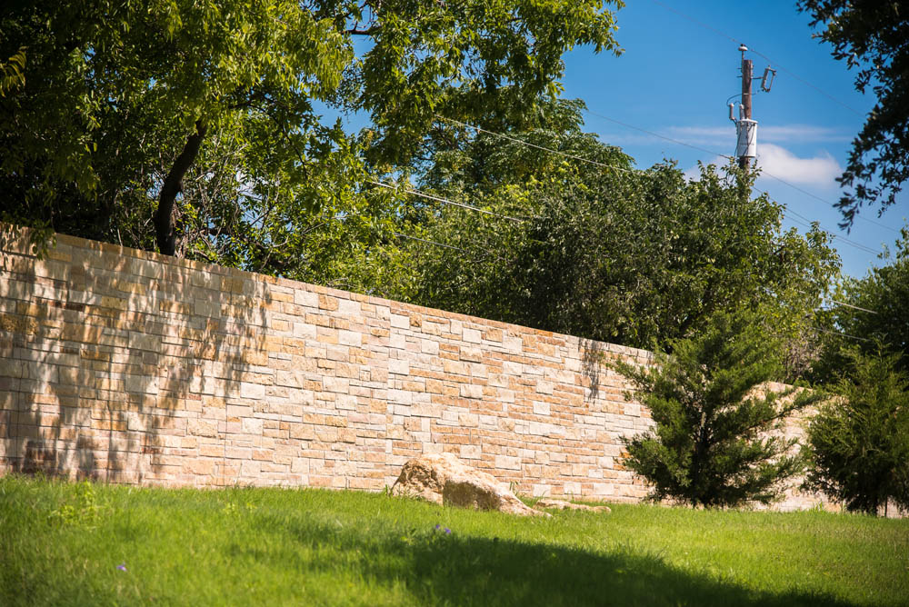 A stone wall runs along a grassy area with trees and bushes on a sunny day. Blue sky and a utility pole are visible in the background.