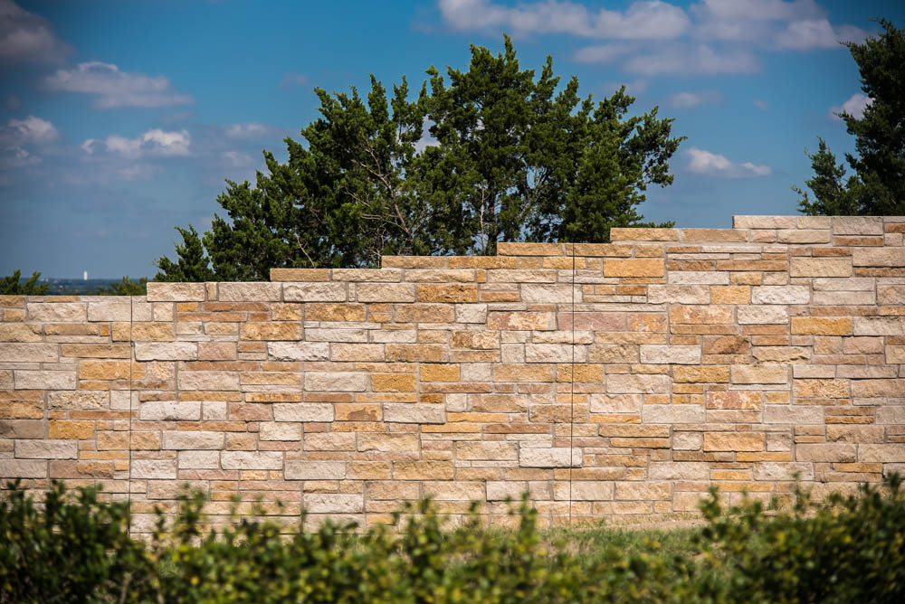 A tan and light brown stone wall stands in front of leafy green trees and bushes under a blue sky with scattered clouds.