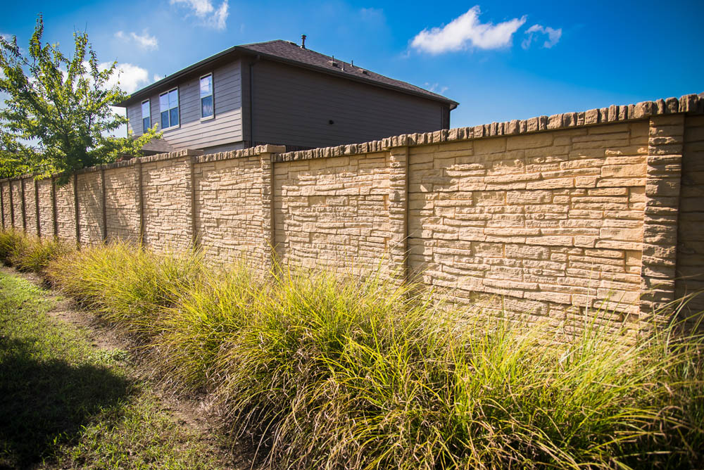 A tall, textured stone fence with vertical columns lines a suburban yard with tall green grass in front, and a house and blue sky with a few clouds in the background.