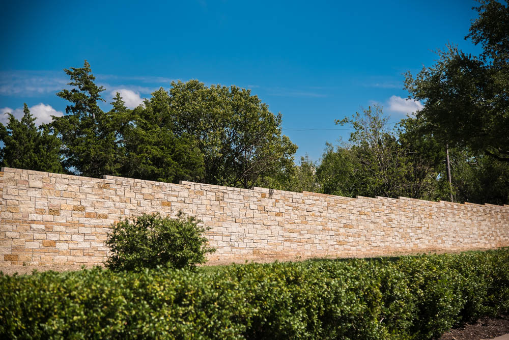 A stone wall runs horizontally with neatly trimmed hedges in front and tall green trees behind it, under a clear blue sky.