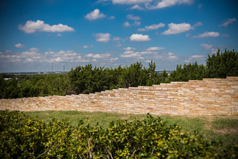 A stone terrace wall with staggered steps is set against green trees and a blue sky with scattered clouds on a sunny day.