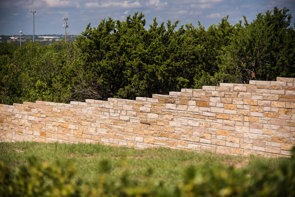 A stone retaining wall with a staggered top edge stands in front of dense green trees under a partly cloudy sky, with grass and shrubbery in the foreground.