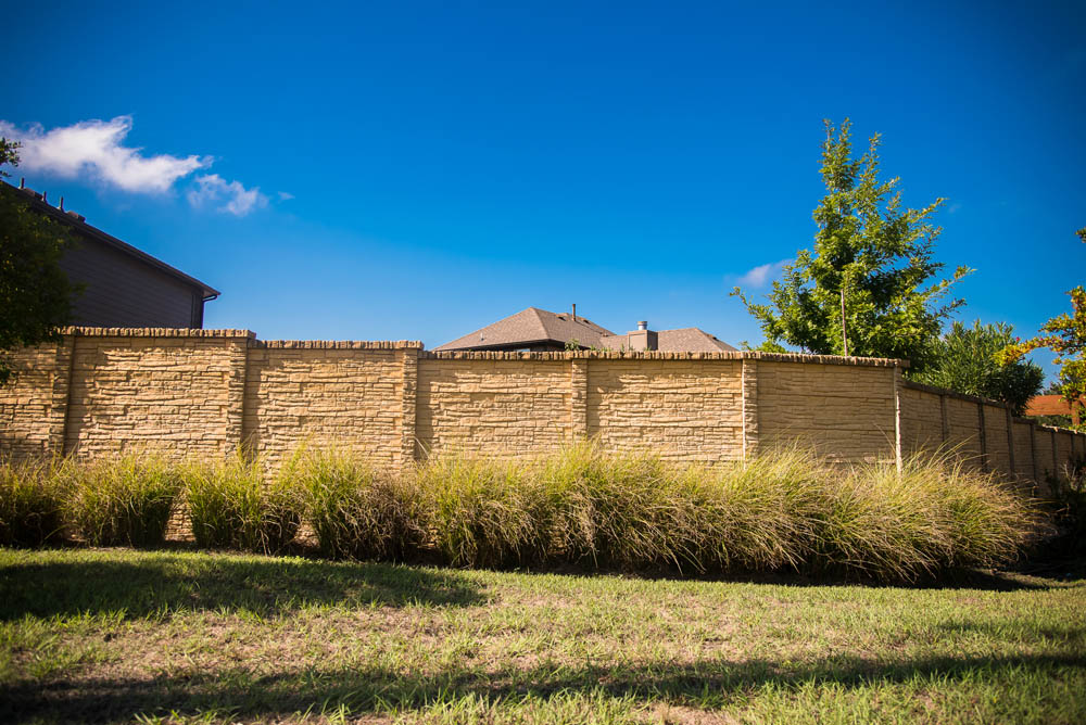 A tall stone fence with decorative brickwork runs along a grassy yard, with tall ornamental grass growing in front of it. Trees and rooftops are visible behind the fence under a clear blue sky.
