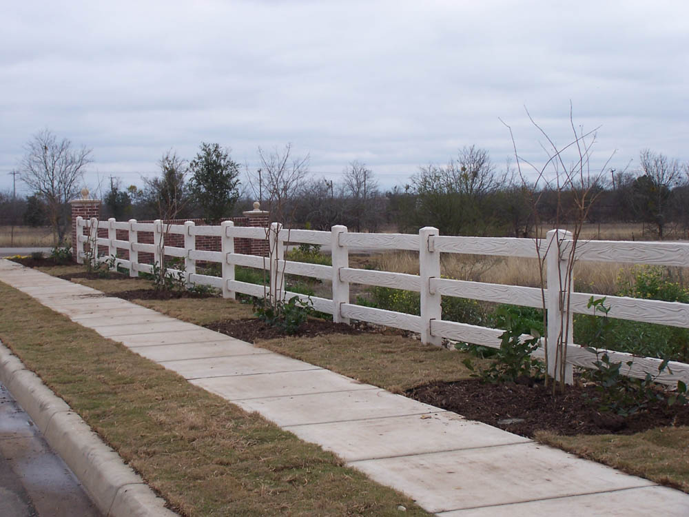 A cement sidewalk runs alongside a grassy area bordered by a white wooden fence, with young plants and bare trees spaced along the edge under a cloudy sky.
