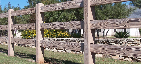 A wooden post-and-rail fence runs alongside a grassy area, with yellow flowers, shrubs, trees, and a stone wall in the background. Houses are partly visible behind the foliage.