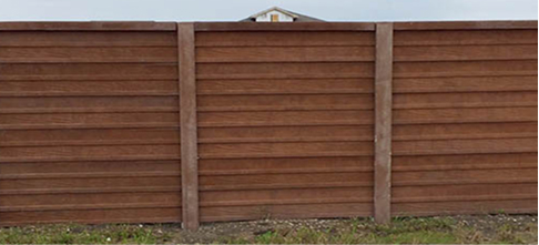 A tall wooden fence with horizontal panels and vertical supports stands in front of a patch of grass and dirt. The roof of a house is just visible above the fence in the background.