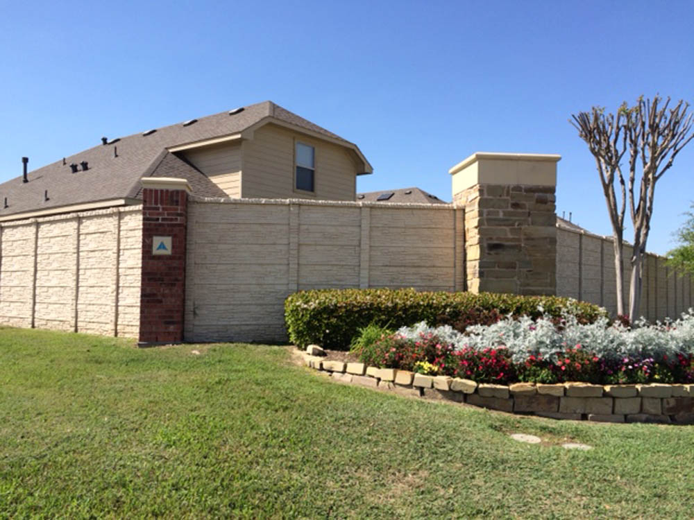 A beige brick privacy wall with brick and stone pillars surrounds a house. In front, there is a landscaped garden with red, white, and green plants bordered by stone, next to a grassy lawn and a pruned tree.