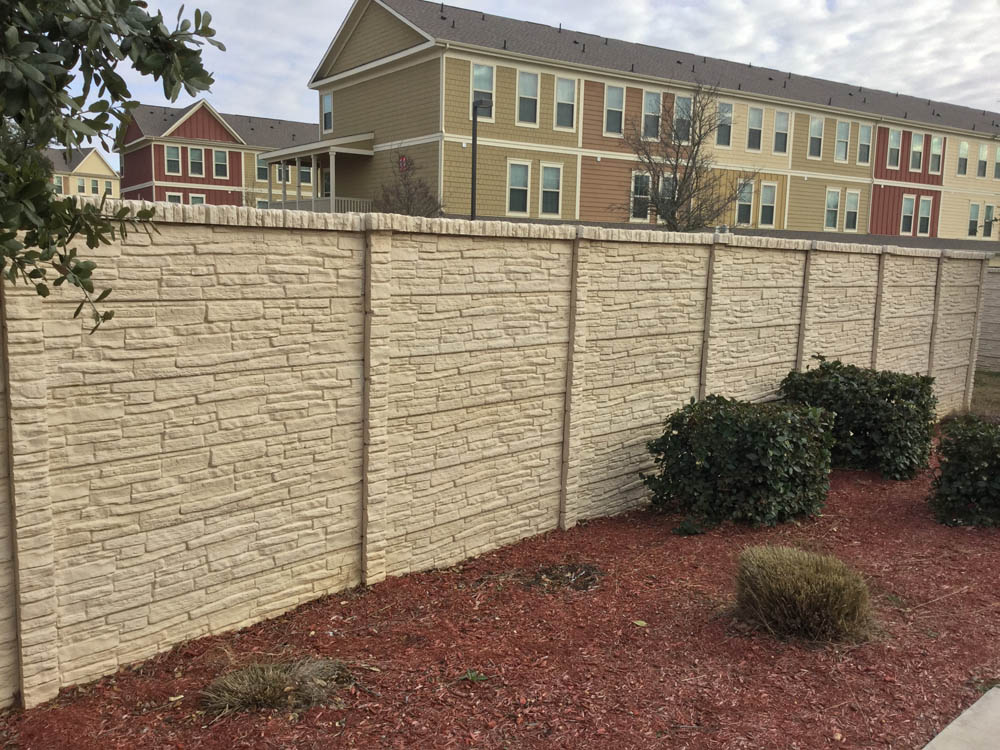 A beige, textured stone-pattern fence stands in front of landscaped bushes and red mulch, with multi-story tan and red apartment buildings visible in the background under a partly cloudy sky.
