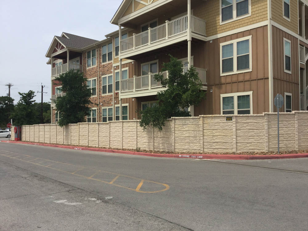 A three-story apartment building with balconies stands behind a tall beige privacy fence along a quiet street with red curbs and yellow parking lines. Small trees and shrubs are visible near the fence.