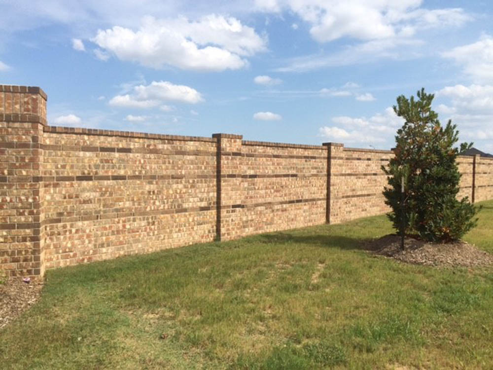 A long brick wall runs across a grassy area under a blue sky with scattered clouds. A small tree is planted in front of the wall to the right.