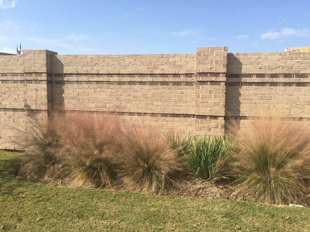 A tall beige brick wall with two horizontal dark lines, standing behind ornamental grasses and green plants growing at its base on a grassy lawn under a blue sky.