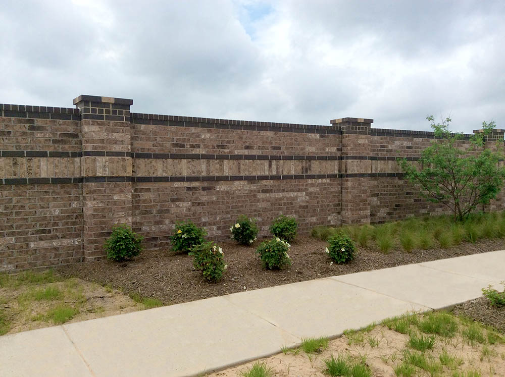 A brick wall with decorative pillars stands behind a row of small bushes and grassy plants, next to a sidewalk under a cloudy sky.