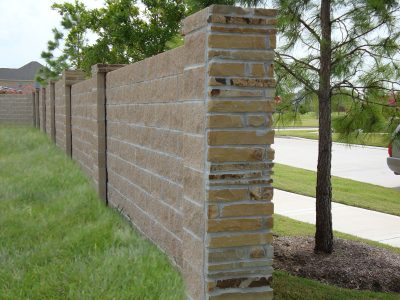 A tall tan stone and brick privacy wall runs along a grassy area, with trees and a parked car visible on the right beside a sidewalk and street.
