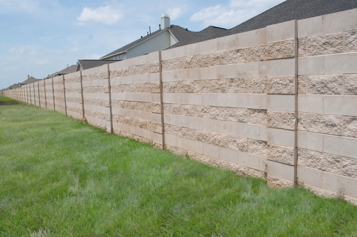 A long beige concrete block wall separates two grassy areas, with houses visible behind the wall and a partly cloudy sky overhead.