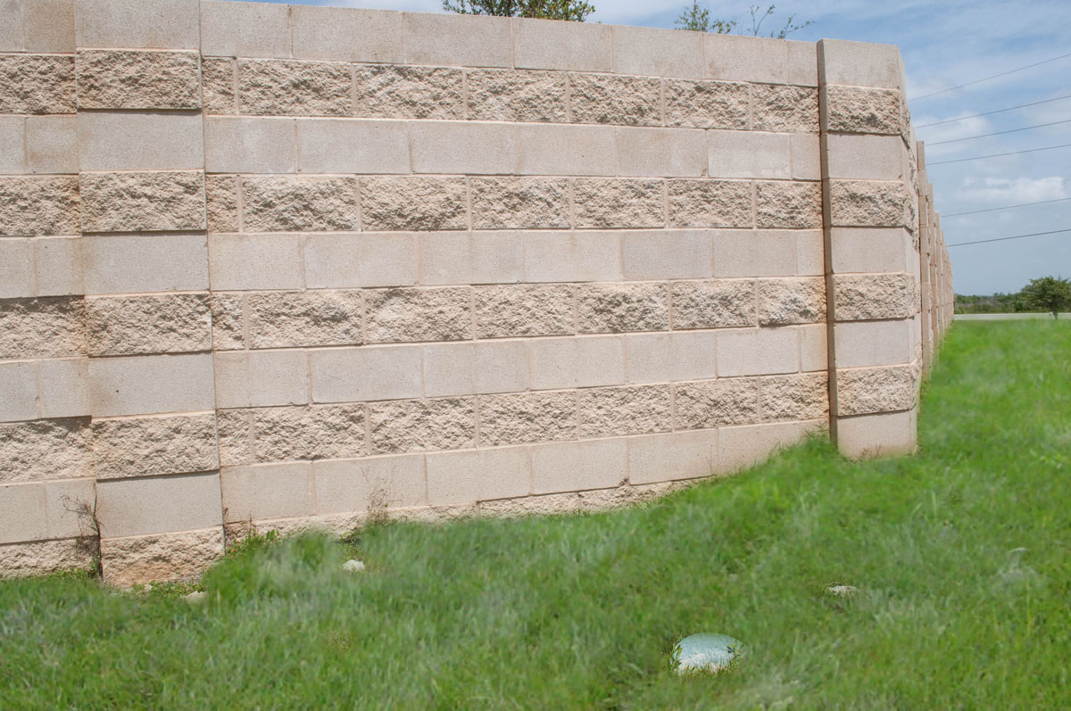 A tan brick wall curves slightly in a grassy area under a blue sky with clouds. There are a few small plants and round objects on the grass near the wall’s base.