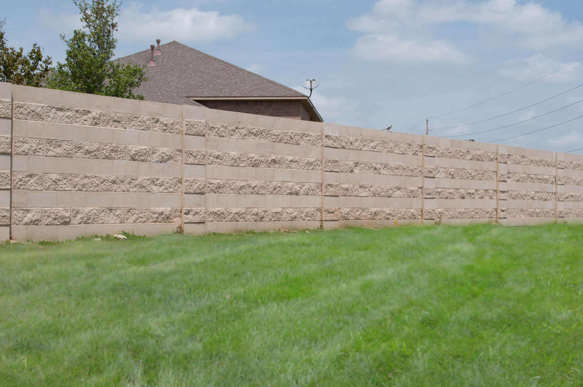 A tall concrete privacy fence with textured horizontal stripes borders a grassy lawn. A house with a brown roof and some trees are partially visible behind the fence under a blue sky with clouds.