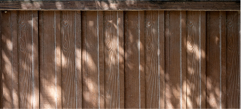 Vertical wooden fence panels with visible wood grain, lightly weathered, and dappled sunlight casting leaf-shaped shadows across the surface.