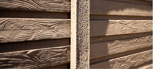 Close-up view of a wooden fence or wall with horizontal planks showing textured grain patterns and a vertical support beam in the center. The wood is painted light brown and appears weathered.