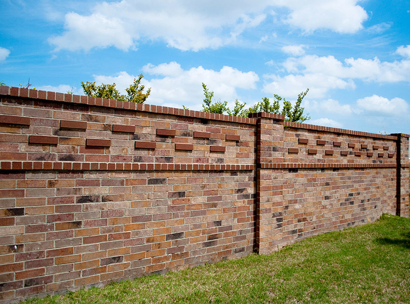 A tall brick wall with a decorative pattern stands next to a grassy area, with green shrubs visible just above the top. The sky is blue and partly cloudy.
