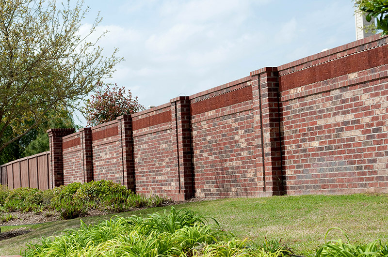 A tall red brick privacy wall runs along a grassy yard with small bushes and trees nearby, under a partly cloudy sky.
