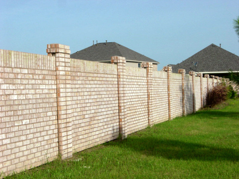 A tall, light-colored brick fence with evenly spaced pillars separates two grassy yards, with houses and a clear blue sky visible in the background.
