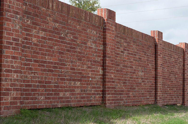 A long, red brick wall with evenly spaced columns stands on grass, extending into the distance under a cloudy sky.