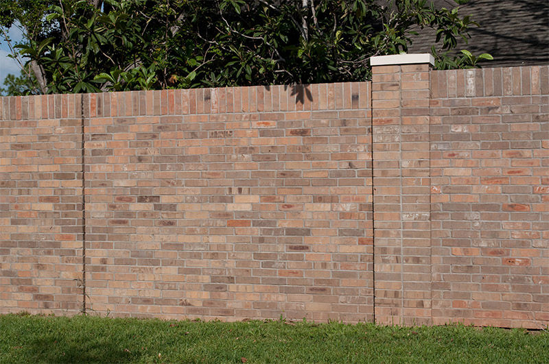 A tall brick wall with a column stands in front of green grass and leafy trees, partially blocking the view of a building behind it.