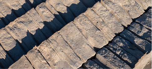 A close-up, aerial view of textured, cracked earth or rock formations with deep grooves and rough, uneven surfaces, highlighted by sunlight and shadow.