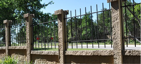 A fence with stone pillars and black iron bars runs alongside a road with green grass and trees in the background under a clear blue sky.