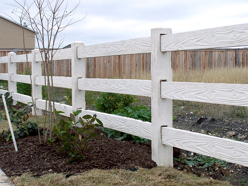 A sturdy white wooden fence with three horizontal rails separates a mulched garden with small plants and a young tree from a grassy and weedy area, with more fencing and a house in the background.
