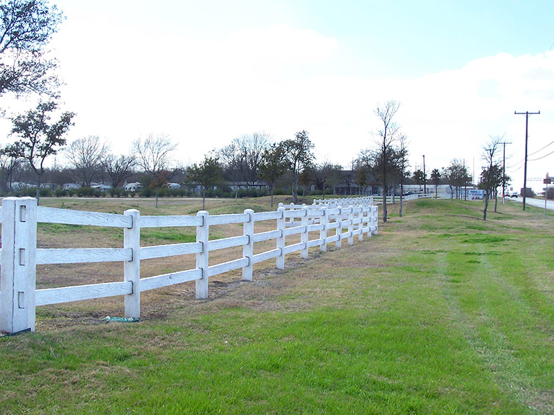 A white wooden fence runs along the edge of a grassy area beside a road with utility poles, trees, and buildings visible in the background under a partly cloudy sky.