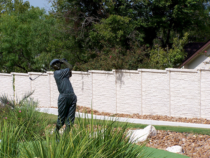 A life-sized statue of a golfer mid-swing stands on a putting green, surrounded by tall grass, rocks, and a white stone fence with trees in the background.