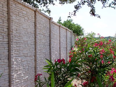 A tall, light-colored stone-patterned wall runs alongside flowering bushes with green leaves and pink blossoms, under a partly cloudy sky with tree branches visible above.