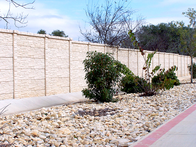 Tan stone privacy wall beside a sidewalk, with a dry landscape of rocks, scattered shrubs, and sparse trees in the background under a partly cloudy sky.