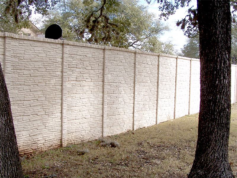 A tall, light-colored concrete fence with a textured, stone-like pattern stands between two large trees in a grassy area. Trees and houses are visible in the background.