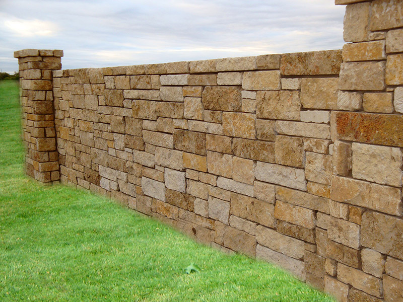 A stone wall with a mix of rectangular and square tan stones runs through a grassy area under a cloudy sky, with two stone pillars at each end.