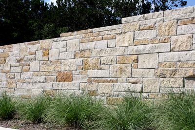 A stone wall made of variously sized beige and tan rectangular blocks stands behind a row of tall green ornamental grasses, with trees visible in the background.