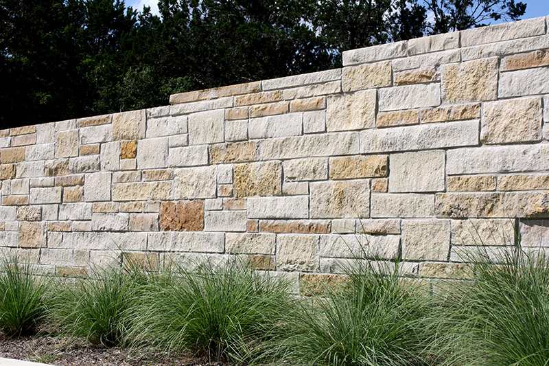A stone wall made of variously sized beige and tan rectangular blocks stands behind a row of tall green ornamental grasses, with trees visible in the background.