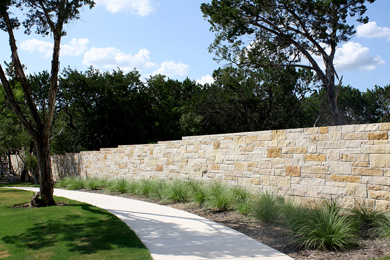 A curved concrete pathway runs alongside a tall stone wall and landscaped grass with small plants, bordered by trees under a partly cloudy sky.
